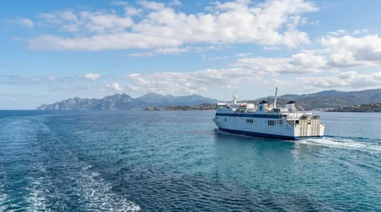 Modernes Fährschiff fährt auf ruhiger See bei hellem Tageslicht, im Hintergrund ist die Küstenlinie Korsikas zu erkennen